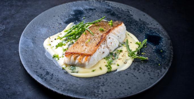 Fotografia um filé de pescada assado sobre um creme de legumes em um prato raso escuro, o qual está sobre uma mesa preta.