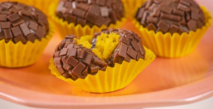 Fotografia em tons de amarelo, rosa e marrom. Ao centro um brigadeiro mordido, mostrando o recheio de bolo de cenoura.