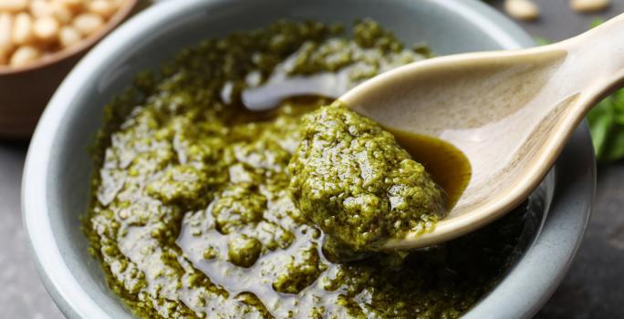 Foto aproximada da receita de molho pesto sem oleaginosas, servido em uma tigela redonda em que há uma colher pegando uma pequena quantidade dele