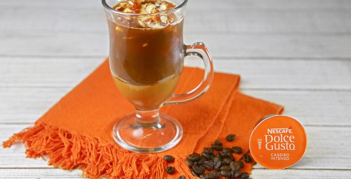 Fotografia em tons de laranja em uma bancada de madeira branca, um pano laranja, uma xícara de vidro com o café com sorvete e pé de moleque dentro dele. Ao lado, uma cápsula de café caseiro intenso Dolce Gusto.