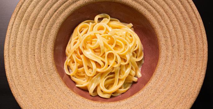 Fotografia em tons de cinza vermelho com um prato vermelho claro ao centro. Dentro do prato existe uma receita de fettuccine Alfredo feita com manteiga e queijo.