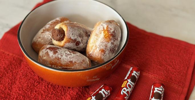 Fotografia mostra um pão tipo bisnaguinha recheado com chocolate baton. Os pãoszinhos estão dentro de um bowl vermelho. Ao lado, batons ao leite dispostos sobre um pano vermelho.