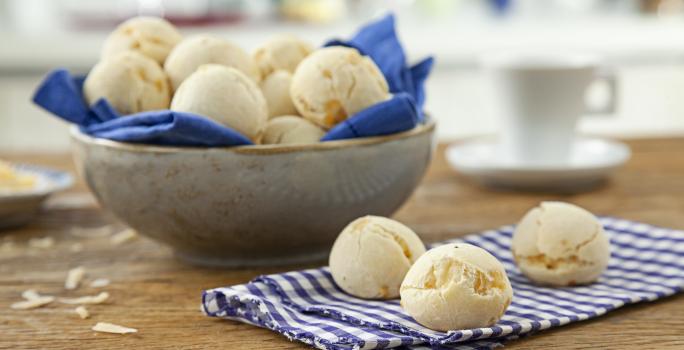 Foto de uma mesa com um pano e 3 pães de queijo redondos em cima. No fundo da foto há uma tigela com mais pães dentro