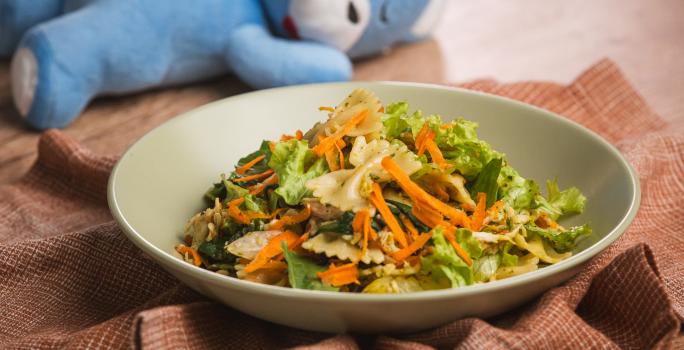 Fotografia em tons de verde e marrom em uma bancada de madeira, um prato redondo fundo verde com a salada de macarrão, frango, legumes e molho pesto dentro. Ao fundo, um ursinho azul deitado.