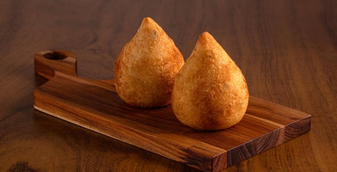 Fotografia de duas coxinhas de frango sobre uma tábua pequena de madeira. A tábua está sobre uma mesa de madeira.