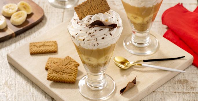 Fotografia em tons de marrom claro com duas taças de vidro ao centro. Dentro de cada taça existe uma receita de banoffee com camadas de biscoito TOSTINES, banana, doce de leite e chantilly.