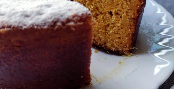 Fotografia em tons de bege em uma bancada com toalha cinza, um prato branco redondo raso, um bolo de milho cortado ao meio com açúcar polvilhado em cima.