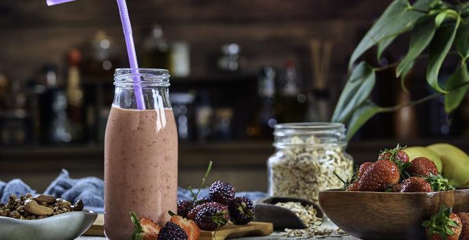 Fotografia de uma bebida densa de morango com leite MOÇA e neston que está em uma garrafa de vidro decorativa, com um canudo roxo e frutas no fundo.