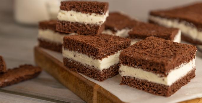 Fotografia de pedaços quadrados de bolo de chocolate recheado com creme de limão, e estão sobre um papel manteiga, apoiado em uma tábua de madeira.