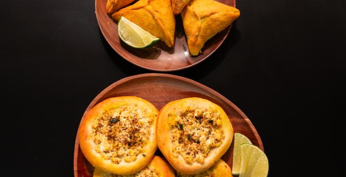 Fotografia em tons de marrom com dois pratos redondos e marrons ao centro. Dentro de um dos pratos existe algumas esfihas fechadas acompanhadas de limão. Dentro de outro prato existe esfihas abertas recheadas de tofu, acompanhadas com limão.