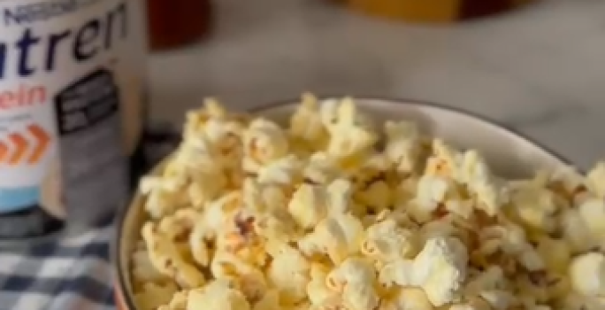 Foto da receita de pipoca doce proteica servida em um bowl laranja sobre uma mesa de mármore branca com um paninho azul xadrez. Ao fundo há uma embalagem de nutren protein e utensílios de madeira.