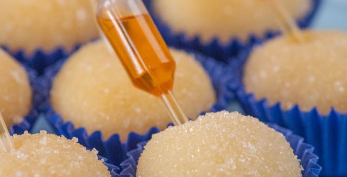 Fotografia de sete forminhas azuis juntas com brigadeiro de pudim, em cada unidade tem uma mini pipeta espetada com calda de caramelo. Os brigadeiros estão apoiados em um apoio decorativo azul claro.