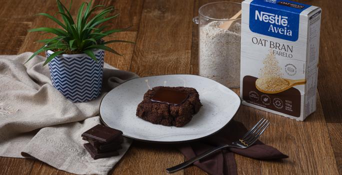 Fotografia de um pedaço de brownie com um quadrado de chocolate ao centro, sobre um prato branco com borda preta. O brownie está ao lado de uma embalagem de aveia farelo, sobre uma mesa de madeira.