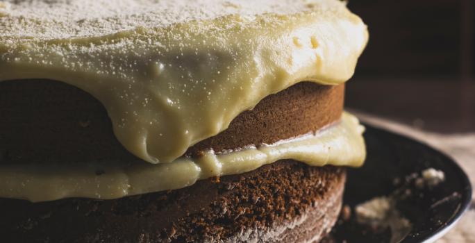 Fotografia de um bolo de abóbora com creme de coco por cima. O bolo está recheado com o mesmo creme, e está sobre um prato raso preto.