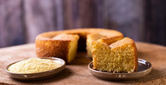 Foto da receita de bolo de milho de liquidificador, redondo, dourado, com uma fatia mais à frente em um prato e ao lado um punhado de fubá em outro prato. Ao fundo é possível ver o bolo inteiro