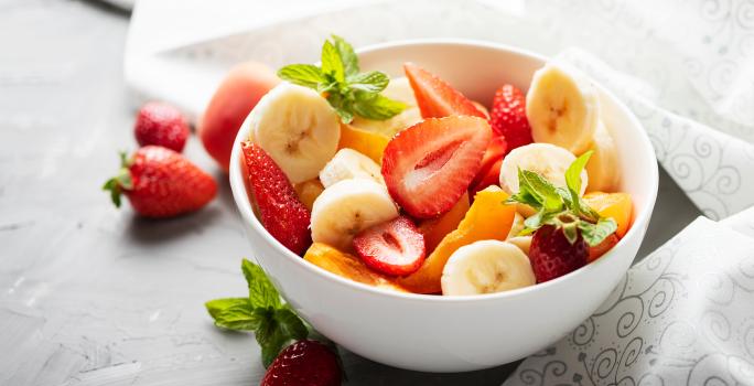 Fotografia de um recipiente pequeno e branco de frutas sobre um purê de banana e folhas de hortelã. O recipiente está sobre uma bancada de cor escura, do lado das frutas tem alguns morangos e mais folhas de hortelã.