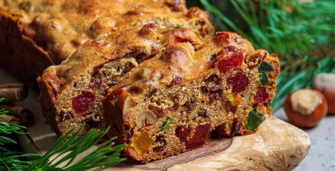 Fotografia de um bolo de frutas cristalizadas e secas com whisky e especiarias, feito na forma pequena de bolo inglês. Tem duas fatias cortadas na frente do restante do bolo, que está sobre um apoio de madeira, ao lado de paus de canela.