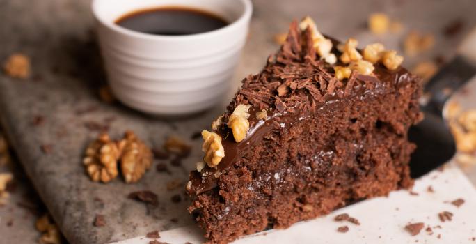 Fotografia de uma fatia de bolo feito com Chocolate NESTLÉ CLASSIC Meio Amargo, nozes e Cobertura de Chocolate ao Leite GAROTO, com raspas de chocolate e castanhas por cima. Ao lado da fatia tem uma xícara pequena branca com café.