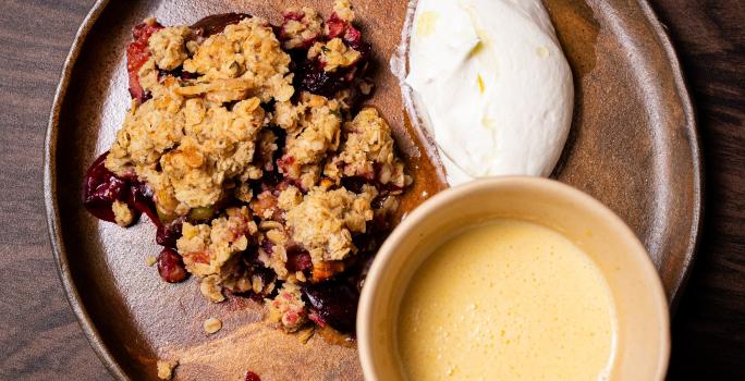 Fotografia vista de cima de um prato raso de vidro marrom, com um crumble de de frutas com creme inglês em um recipiente e chantilly de iogurte ao lado. O prato está sobre uma mesa de madeira.