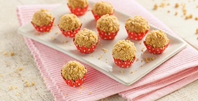 Fotografia em tons de branco e vermelho de bancada de madeira branca, pano rosa dobrado, um brigadeiro de paçoca sobre o pano,  prato retangular branco com nove brigadeiros de paçoca em forminhas vermelhas com bolinhas brancas.