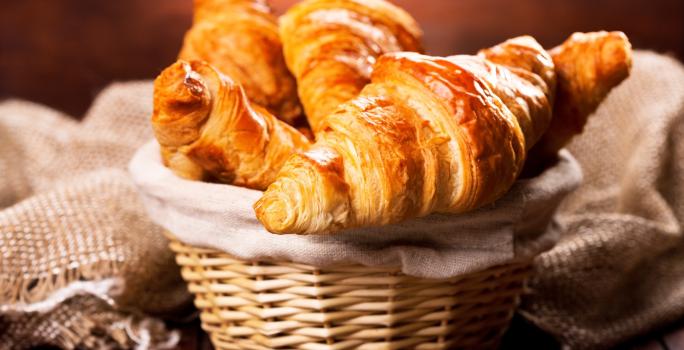 Fotografia de cestinhas de palha com um pano de tom bege. Dentro da cestinha no centro da foto, tem vários pães tipo croissant.