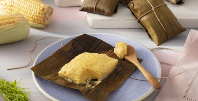 Foto da receita de Pamonha de Leite Moça, Milho Verde e Erva Doce. Observa-se a pamonha aberta em um prato de sobremesa de cerâmica com uma colherinha ao lado direito. Atrás, tem milhos na espiga e pamonhas fechadas e embrulhadas para decorar.