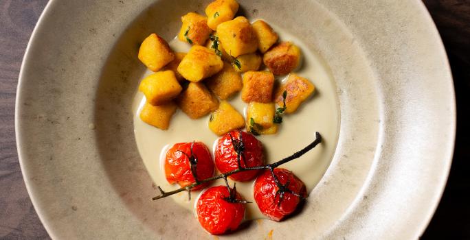 Fotografia em tons de branco com um prato branco ao centro. Dentro do prato existe uma receita de nhoque de abóbora acompanhada de tomates confitados