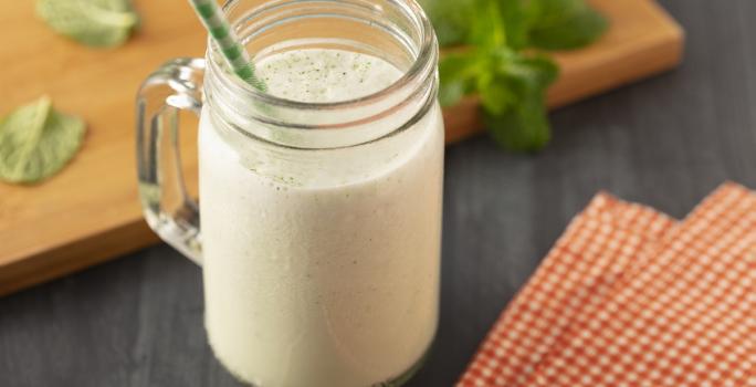 Fotografia em tons de branco, em uma bancada de madeira de cor preta. Ao centro, um copo com o suco de melão com um canudo verde dentro. Ao lado do copo, há um pano quadriculado e ao fundo, uma tábua de madeira de cor marrom folhas de hortelã espalhadas.