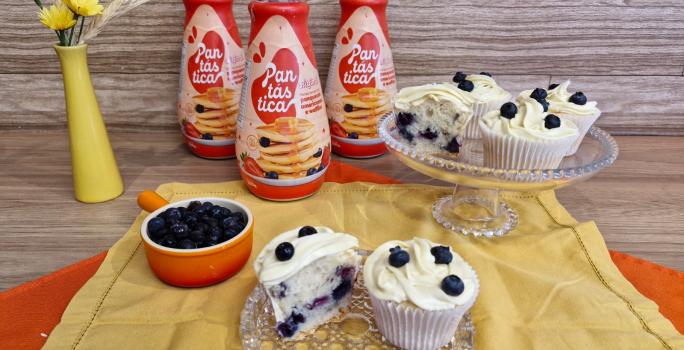 Fotografia em tons de amarelo com dois recipientes de vidro ao centro. Em cima dos recipientes existe vários cupcakes recheados com blueberry e coberto com um creme branco feito de GALAK.