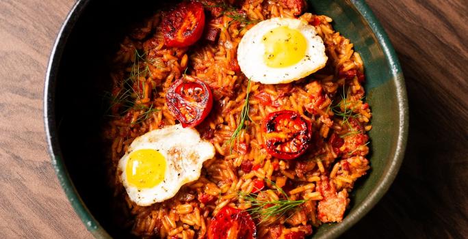 Fotografia em tons de marrom com um recipiente fundo e preto ao centro. Dentro do recipiente existe uma receita de arroz com linguiças. Em cima existe dois ovos de codorna fritos e tomates cereja tostados na frigideira.