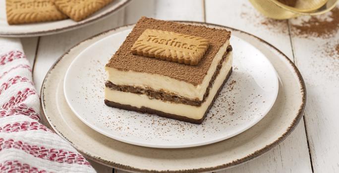 Foto da receita de Tiramisú de Tostines Especiarias. Observa-se uma fatia do Tiramisú bem alta expondo as camadas de creme e de biscoito. No topo, observa-se cacau em pó polvilhado com um Biscoito Tostines.