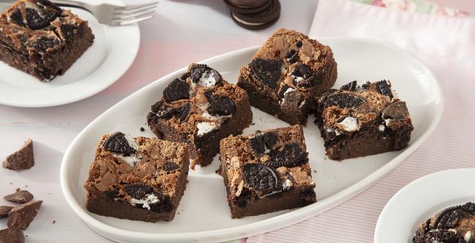 Foto da receita de Brownie de Leite Moça e Biscoito Negresco. Observa-se 7 fatias de brownie cortadas e dispostas em uma forma branca oval em um fundo rosa pastel.