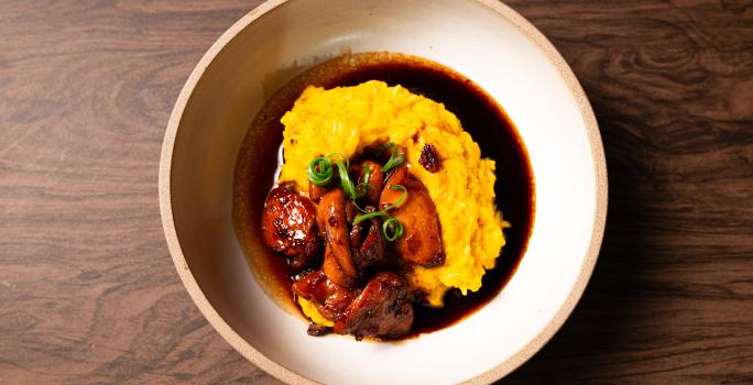 Fotografia vista de cima de uma receita com arroz cremoso na cor amarela sobre de um molho com shoyu. Por cima do arroz tem uns pedaços de frango caramelizados, e cebolinha para enfeitar.