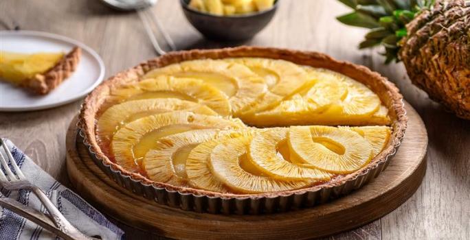 imagem de uma torta redonda com rodelas de abacaxi assadas por cima. A torta permanece em uma forma de tortas com as laterais chanfradas e está sobre uma tábua de mandeira. ao fundo, um pedaço da torta fica sobre um prato branco e ao lado um abacaxi.