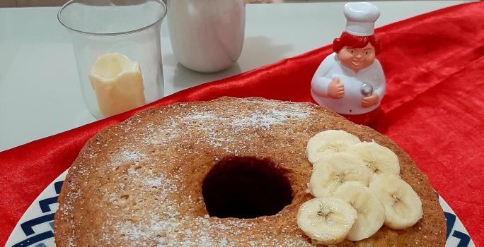 Fotografia em tons de azul em uma mesa com um pano vermelho, um prato redondo raso com desenho em azul e um bolo de banana com canela e rodelas de banana em cima dele.