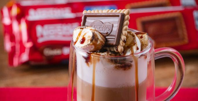 Fotografia em tons de vermelho com uma caneca de vidro ao centro. Dentro da caneca existe uma receita de café com leite finalizada com marshmallow e Choco Biscuit NESTLÉ