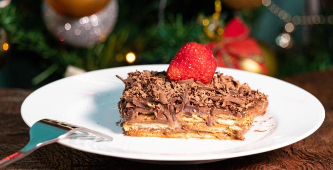 Fotografia de um pedaço de pavê de damasco com chocolate, cobertura de ganache, raspas de chocolate e um morango por cima. O pavê está em um prato raso branco, o qual está sobre uma mesa de madeira.