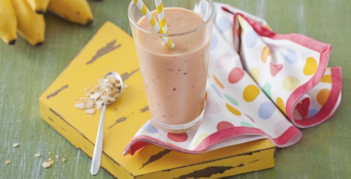 Fotografia em tons de verde e amarelo de uma bancada verde com uma tábua amarela, sobre ela um paninho com bolinhas coloridas e um copo com vitamina de frutas. Ao lado uma colher. Ao fundo um cacho de banana.