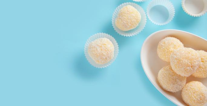 Fotografia vista de cima de cinco docinhos recheados com geleia de damasco e passados no coco dentro de um recipiente oval e fundo de cor branca. Ao lado tem quatro forminhas de brigadeiro, e duas estão com o docinho, e estão sobre uma mesa azul.