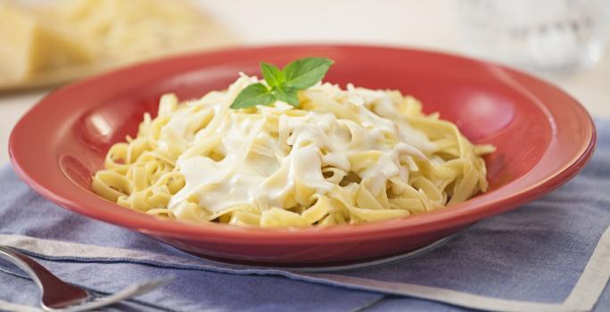 Fotografia em tons de vermelho e azul em uma bancada de madeira com jogos americanos azul e um prato vermelho com o macarrão ao molho branco dentro. Ao fundo, uma peça de queijo parmesão em fatias.