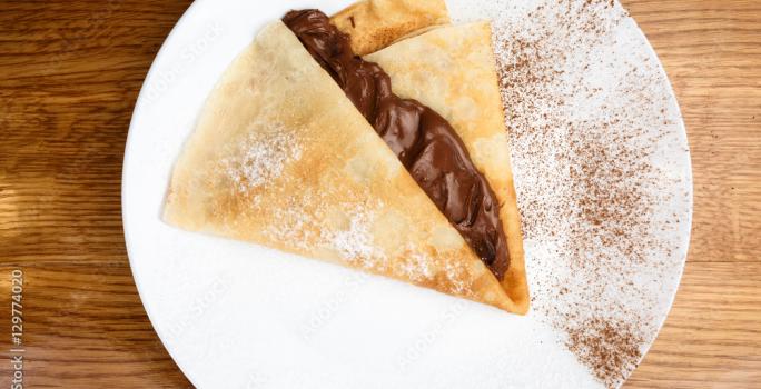 Fotografia vista de cima de uma panqueca com calda quente de mel e chocolate. A panqueca está em um prato branco raso com um pouco de cacau em pó peneirado por cima. O prato está sobre uma mesa de madeira.