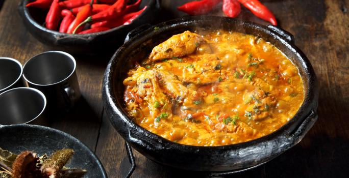 Fotografia de uma panela de barro com caldeirada borbulhando de peixe com cação, postas de pescada e corvina, molho de tomate, açafrão, louro e salsinha.