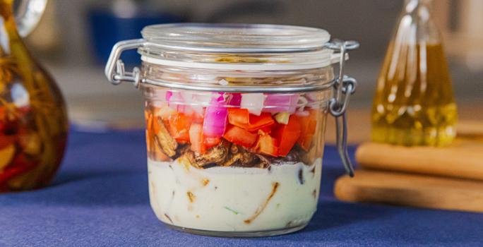 Fotografia em tons de azul com um pote de vidro ao centro. Dentro do pote existe uma salada de berinjela assada com molho de iogurte, tomate e cebola roxa