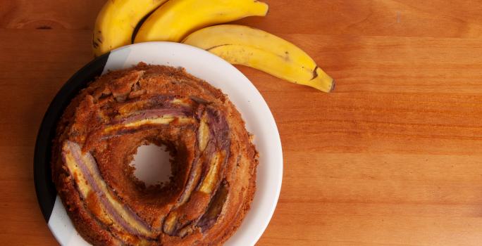 Fotografia em tons de marrom claro com um prato branco ao centro. Dentro do prato existe um bolo de banana redondo com um furo no meio, por cima do bolo existe pedaços de bananas caramelizadas. Ao lado existe três bananas inteiras.