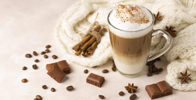 Fotografia em tons de branco de uma xícara de café de vidro com cappuccino feito de café, chocolate em pó, canela e laranja. Por cima do cappuccino tem canela salpicada, ao lado da xícara tem canelas em pau, chocolates, anis estrelado e uma lã branca.