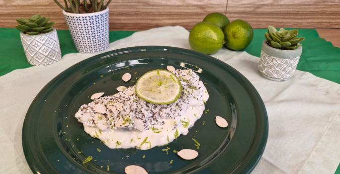 Foto em tons de branco da receita de tapioca servida com uma rodela de limão por cima, sobre um prato preto em cima de uma toalha de linho com limões e vasinhos de planta ao lado