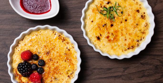 Fotografia vista de cima de dois cremes brulée, um de alho poró com queijo canastra e o outro, sabor cumaru com pistache e frutas vermelhas, os dois cremes brulée estão em ramekins. Ao lado tem uma calda de frutas vermelhas.