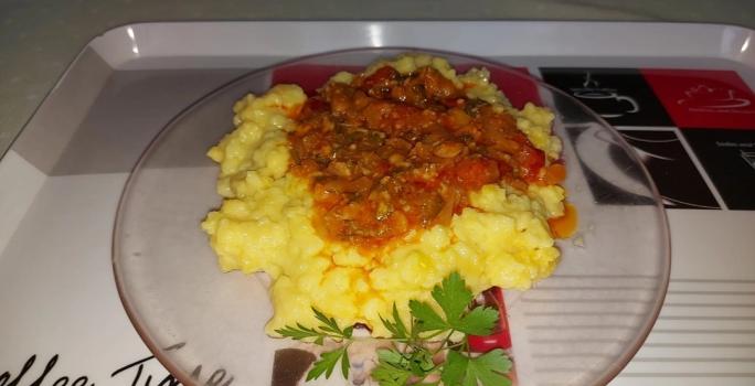 Fotografia de um prato raso transparente com uma polenta caseira. Por cima tem molho de tomate e, ao lado, tem salsinha.