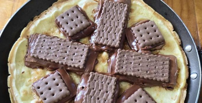 Fotografia em tons de marrom e vermelho com um prato preto ao centro. Em cima do prato tem uma pizza feita de massa de ovo e tapioca. Por cima da massa existe vários biscoitos Choco Trio derretidos.