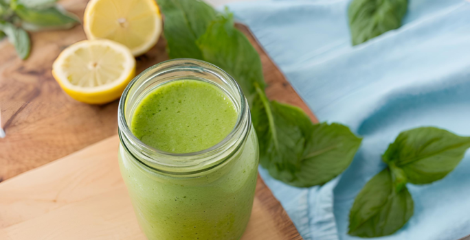 Fotografia de um copo de vidro decorativo com suco detox de cor verde feito com couve, maçã verde, pepino, gengibre e nutren. O copo está sobre uma tábua de madeira, ao lado de folhas de hortelã e metades de limão siciliano sobre a mesa.